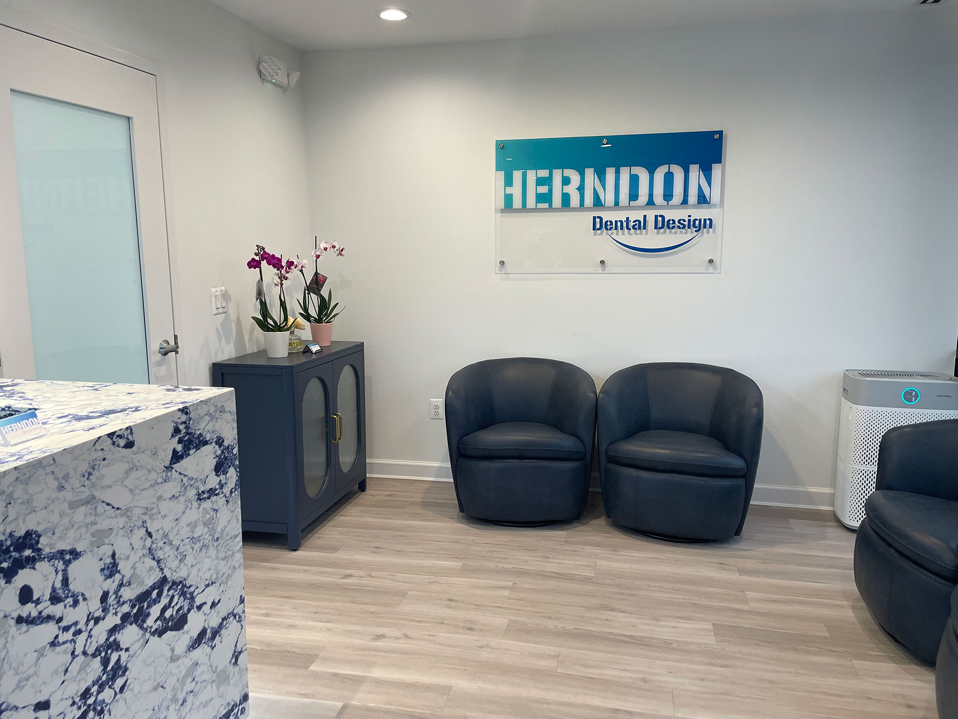 An interior office space with a modern reception area featuring a marble countertop, black leather chairs, a white sign with blue lettering that reads Hermondon Dental Design, and a large window letting in natural light.